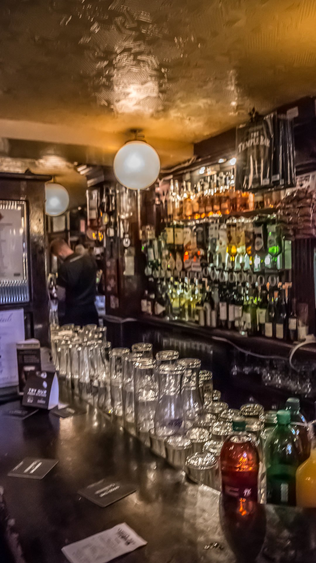 A dim pub bar in Temple Bar, with bottles, glasses, and round white lamps.