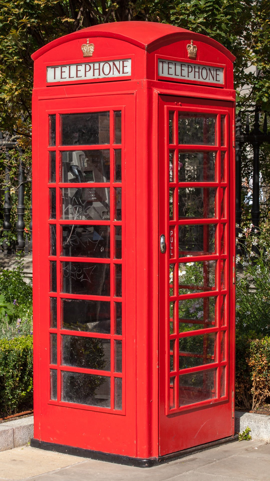 A bright red British telephone box, seen straight on, with trees behind it.